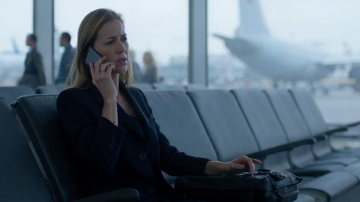 A frustrated businesswoman on her phone sits in an airport gate seat with her hand on her carry-on bag, an airliner outside the window.