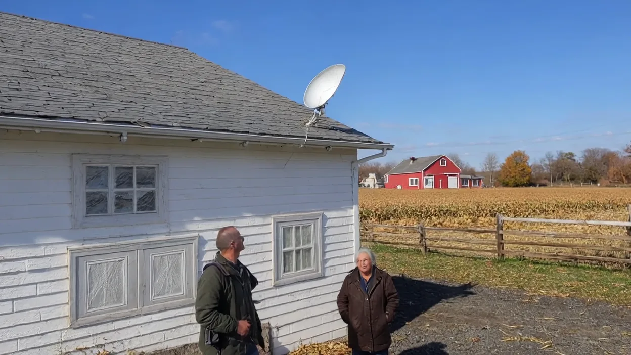An installer and an older woman look up at a satellite dish mounted on the roof of a white farmhouse, a red barn and cornfield behind them.
